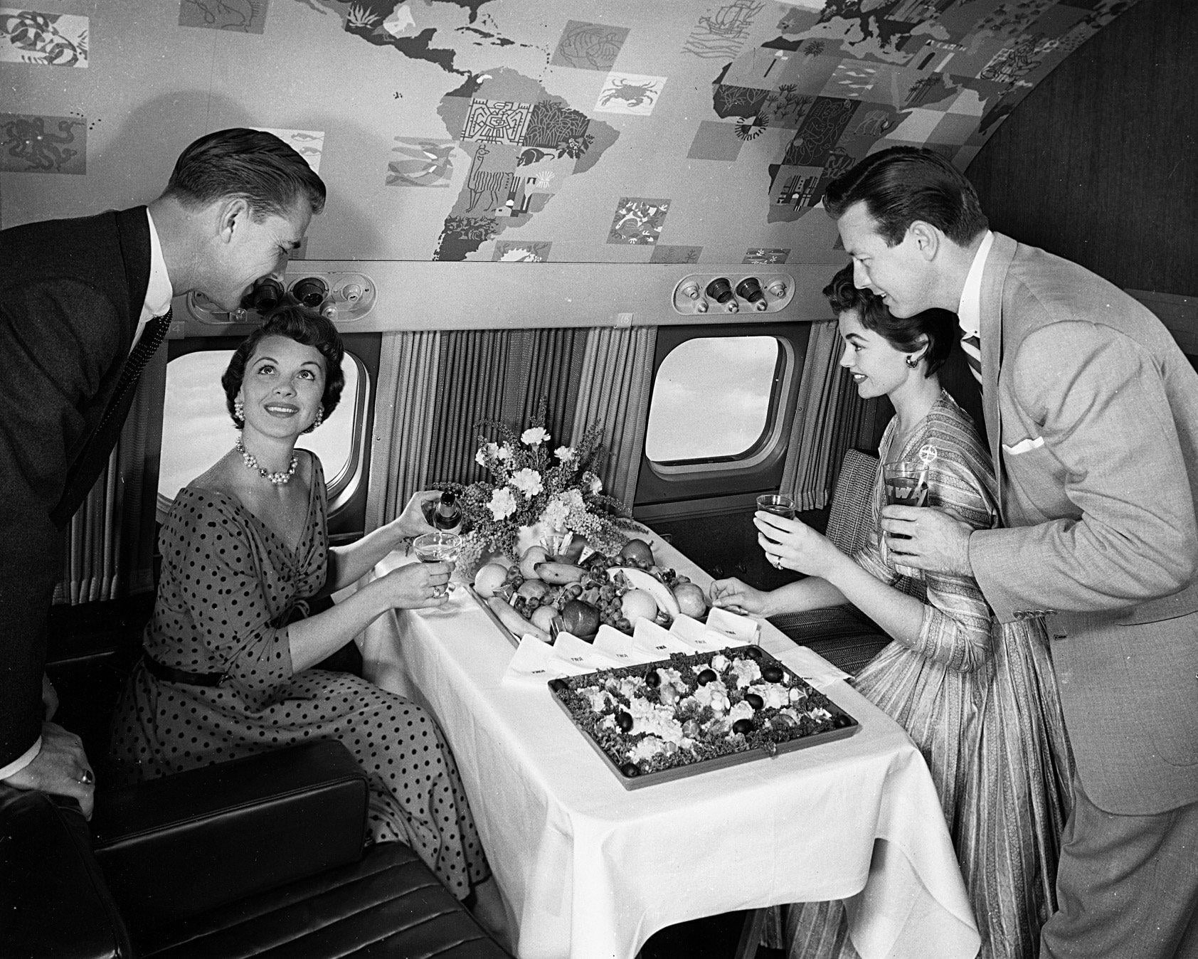 Four well-dressed adults enjoy a meal and drinks around a table with fruit and flowers inside an airplane cabin with decorated ceiling panels.