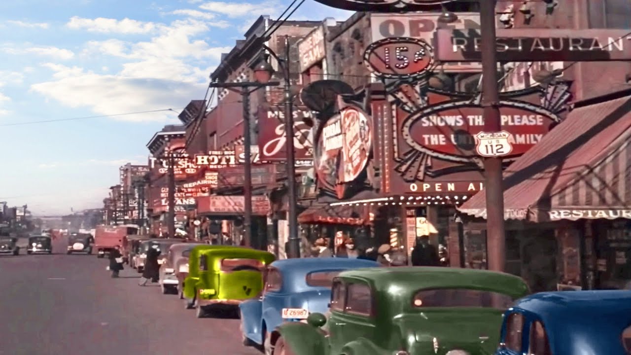 A busy street from the 1940s lined with colorful vintage cars and buildings featuring bright neon signs for restaurants, bars, and entertainment venues under a partly cloudy sky.