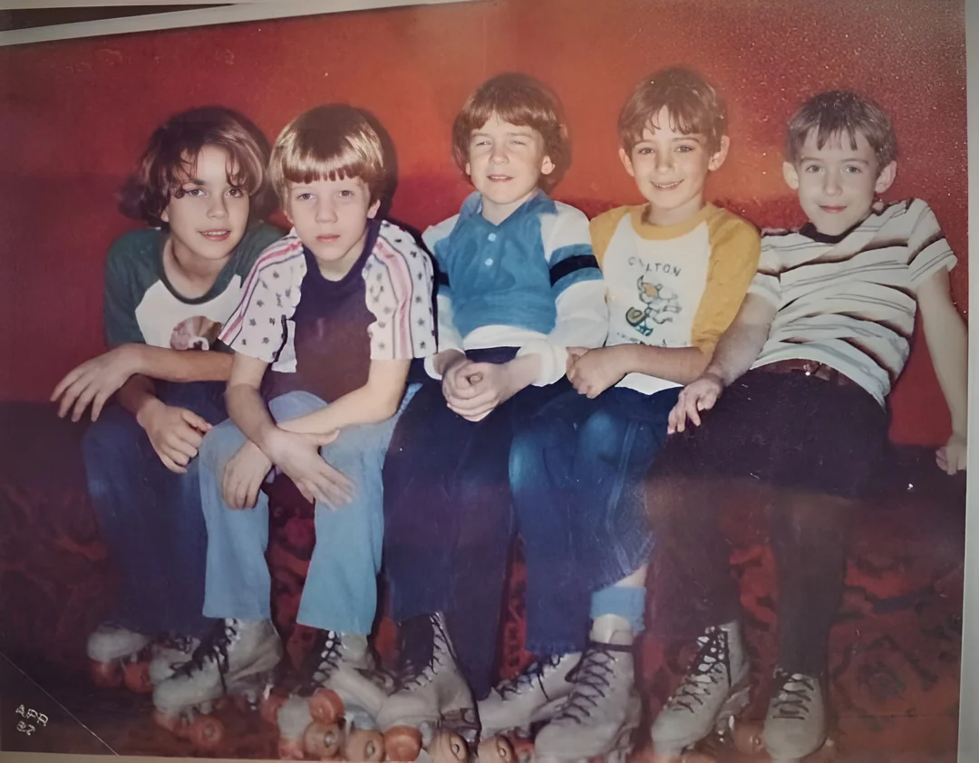 Five children wearing roller skates sit closely on a bench against a red wall, smiling at the camera. They are dressed in colorful, casual 1970s-style clothing.
