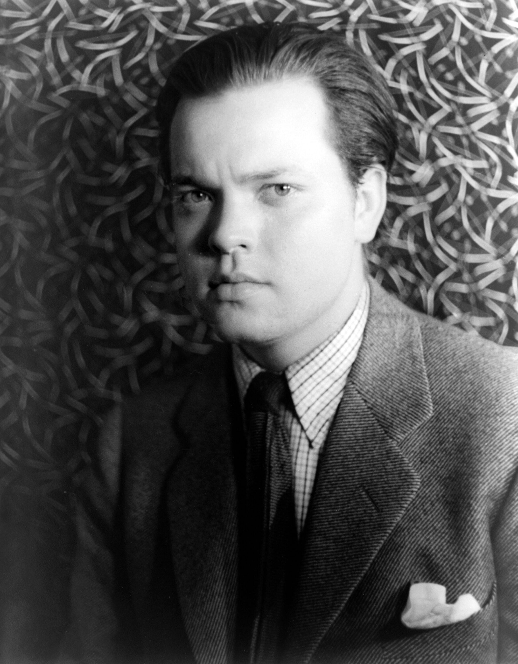 A young man in a suit and tie looks seriously at the camera, with a patterned background behind him.