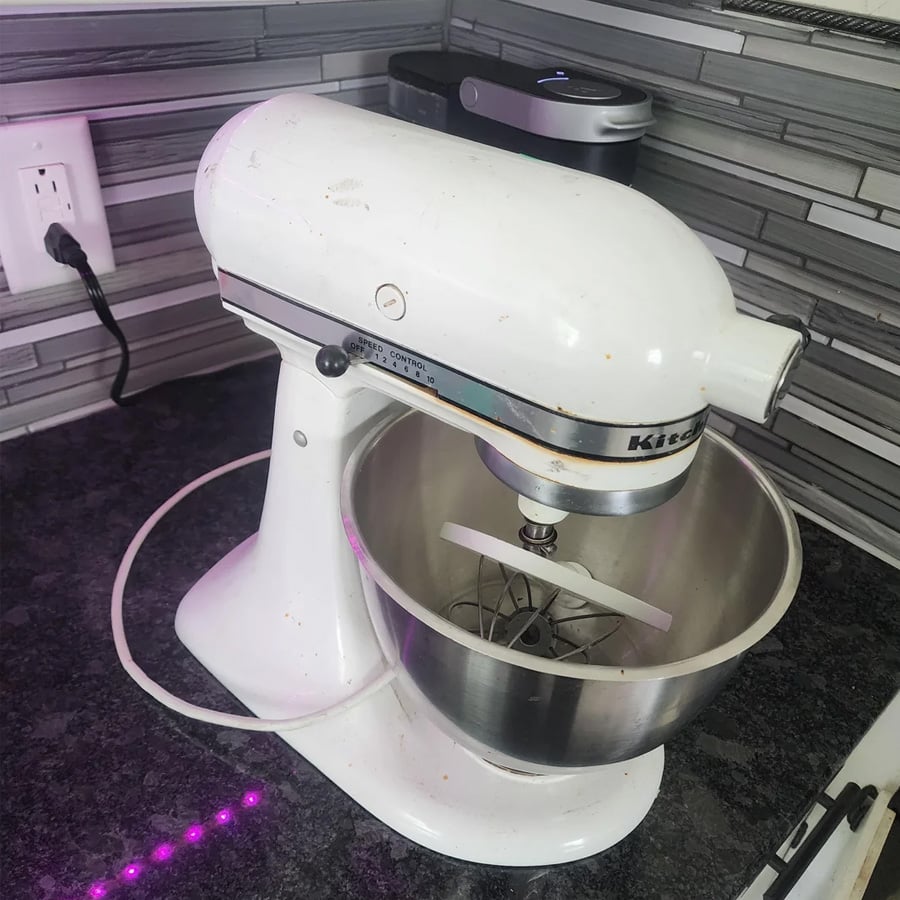 A white stand mixer with a metal mixing bowl sits on a dark granite countertop in a modern kitchen with gray tiled backsplash.