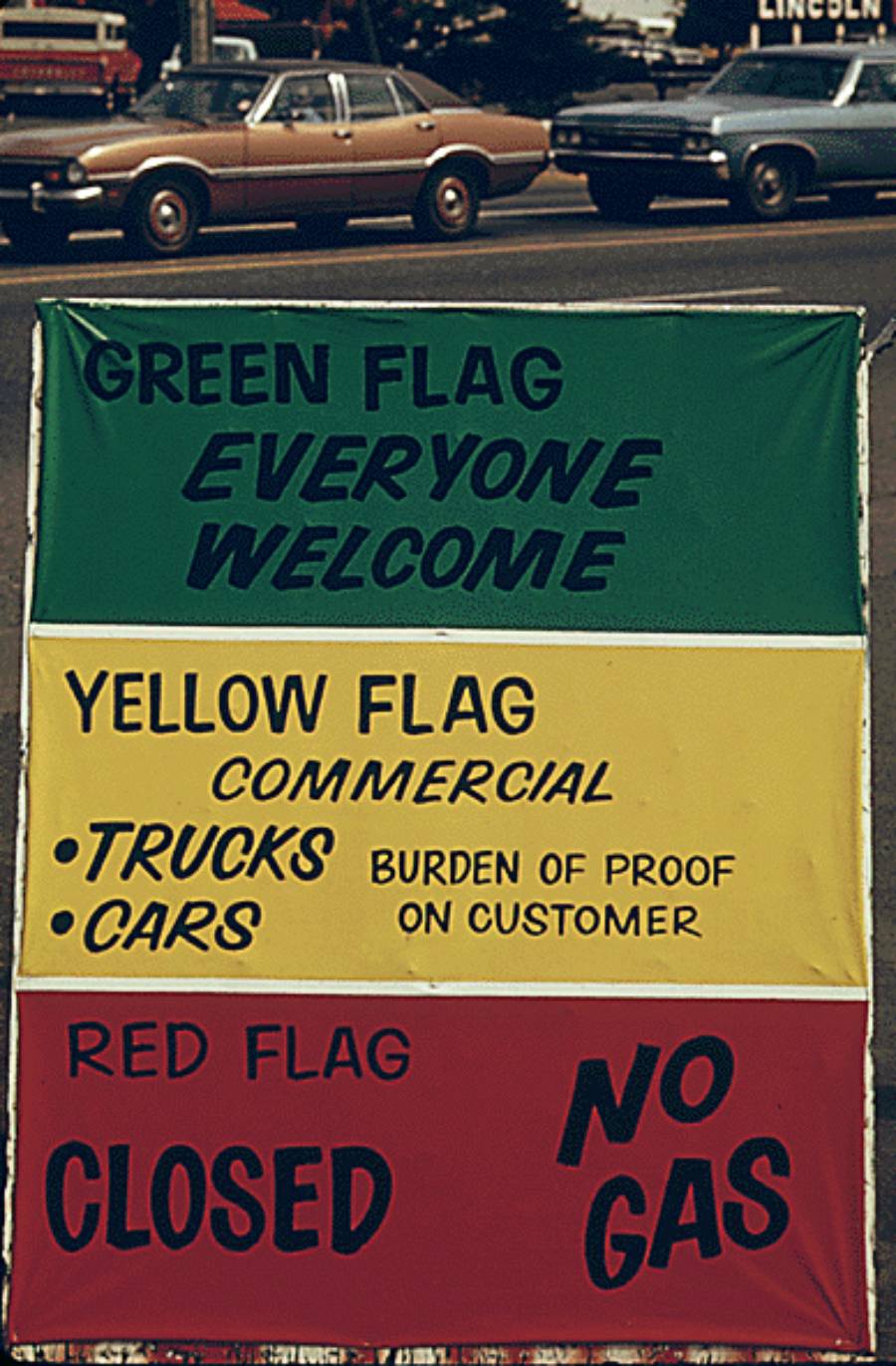 A sign with green, yellow, and red sections outlines gas station policies: green says “Everyone Welcome,” yellow details commercial truck and car services, and red indicates “Closed, No Gas.” Cars are parked in the background.