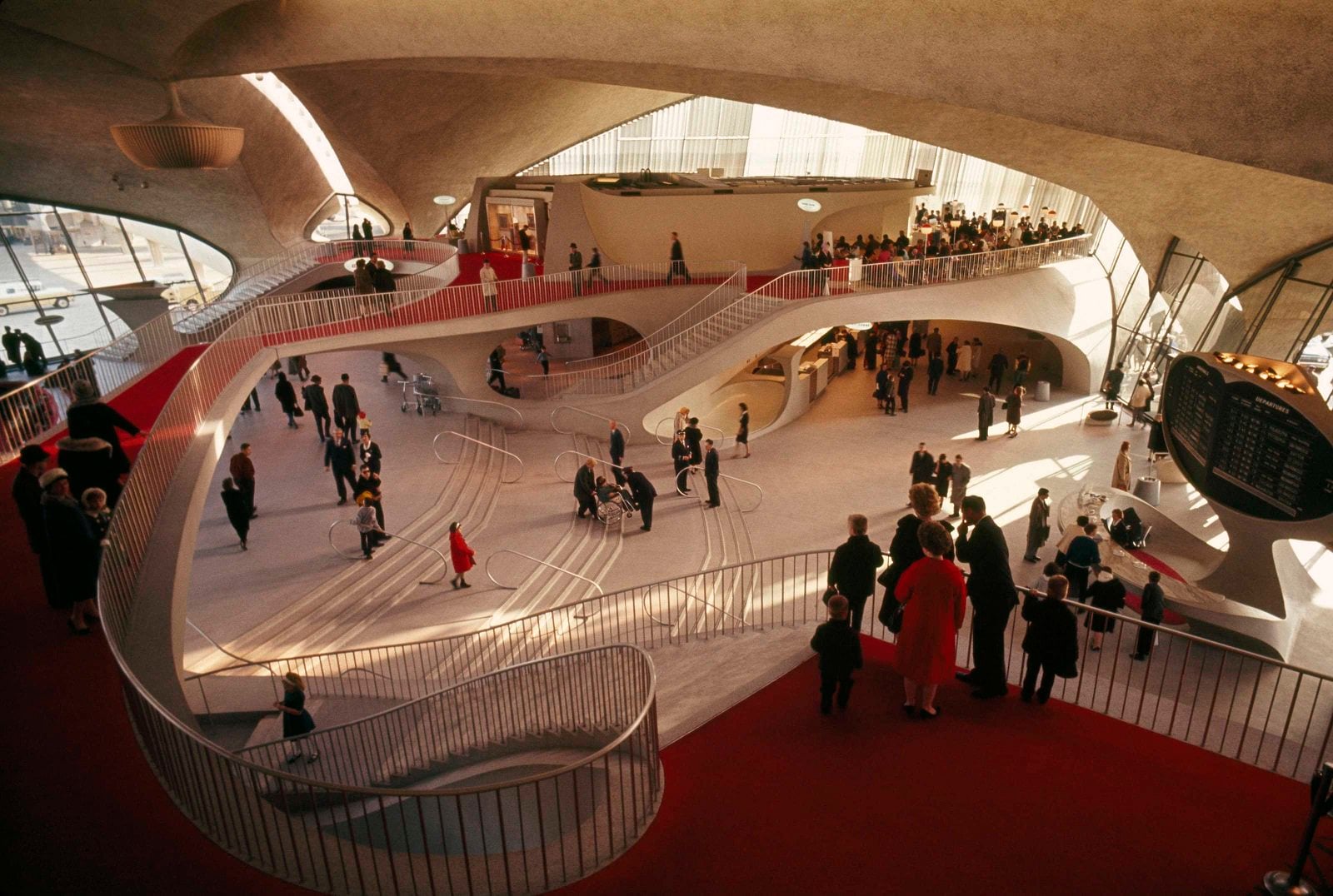 People gather and walk inside a modern, curvy building with sweeping staircases, red carpets, and large windows, likely an airport terminal, featuring a departure board and stylish mid-century design elements.