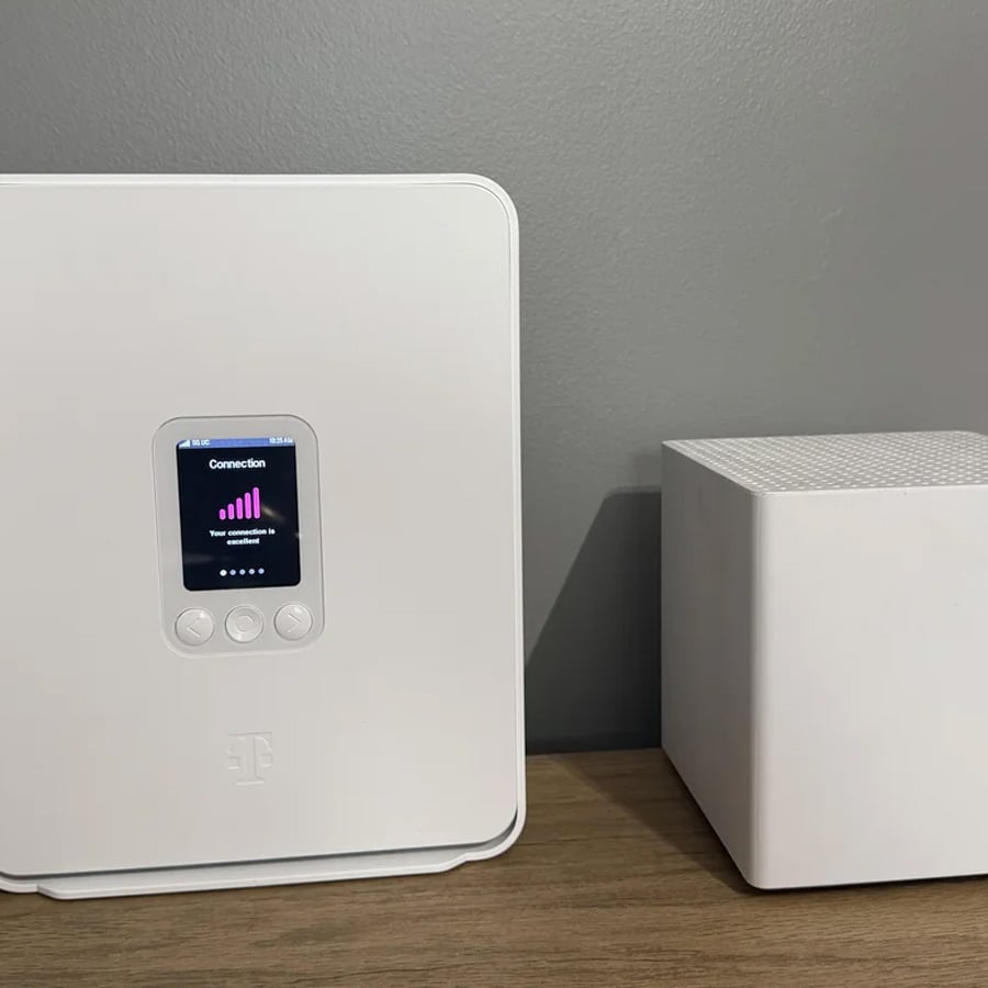 A white wireless internet gateway with a small display showing signal strength sits next to a white mesh network cube on a wooden surface against a gray wall.