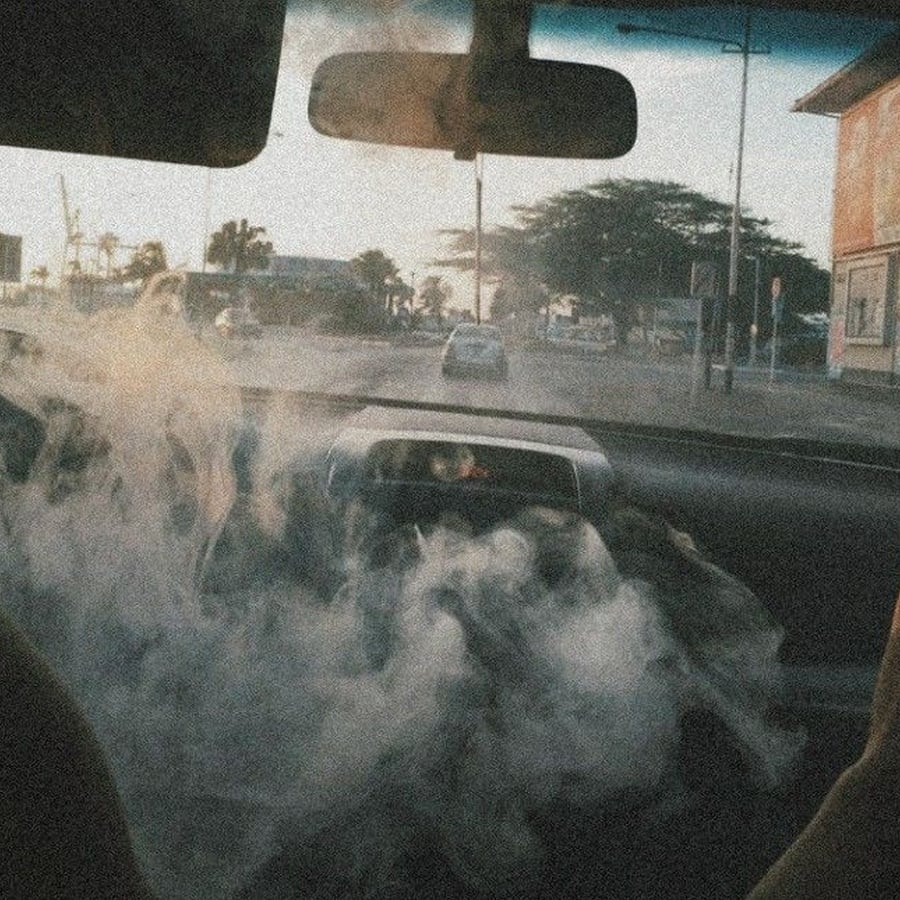 A car interior filled with smoke, looking out through the windshield at a street with trees, buildings, and a few cars in the distance during daylight.