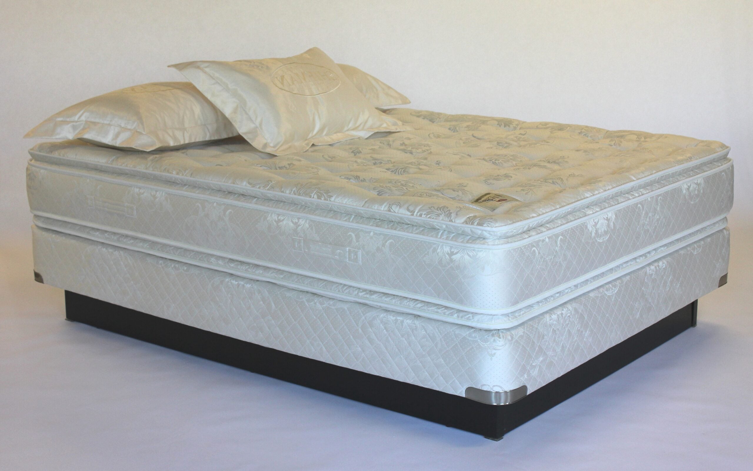 A neatly made bed with two pillows and a thick, patterned mattress on a black platform base against a plain background.