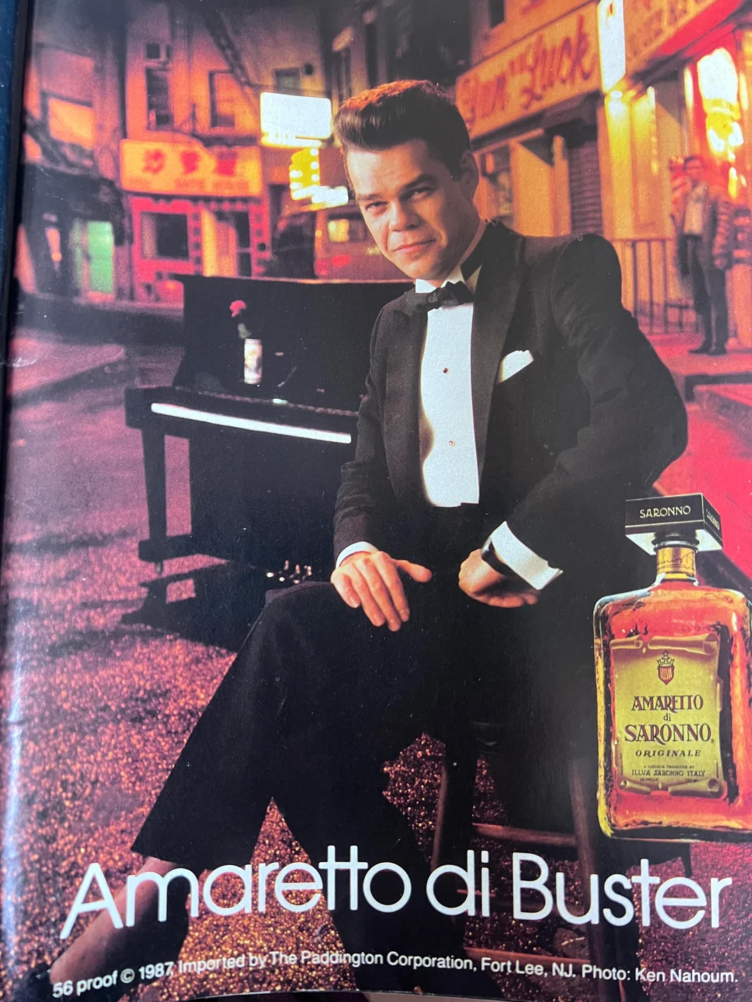 A man in a tuxedo sits by a piano on a city street at night. A bottle of Amaretto di Saronno liqueur is in the foreground. The text reads, “Amaretto di Buster.”