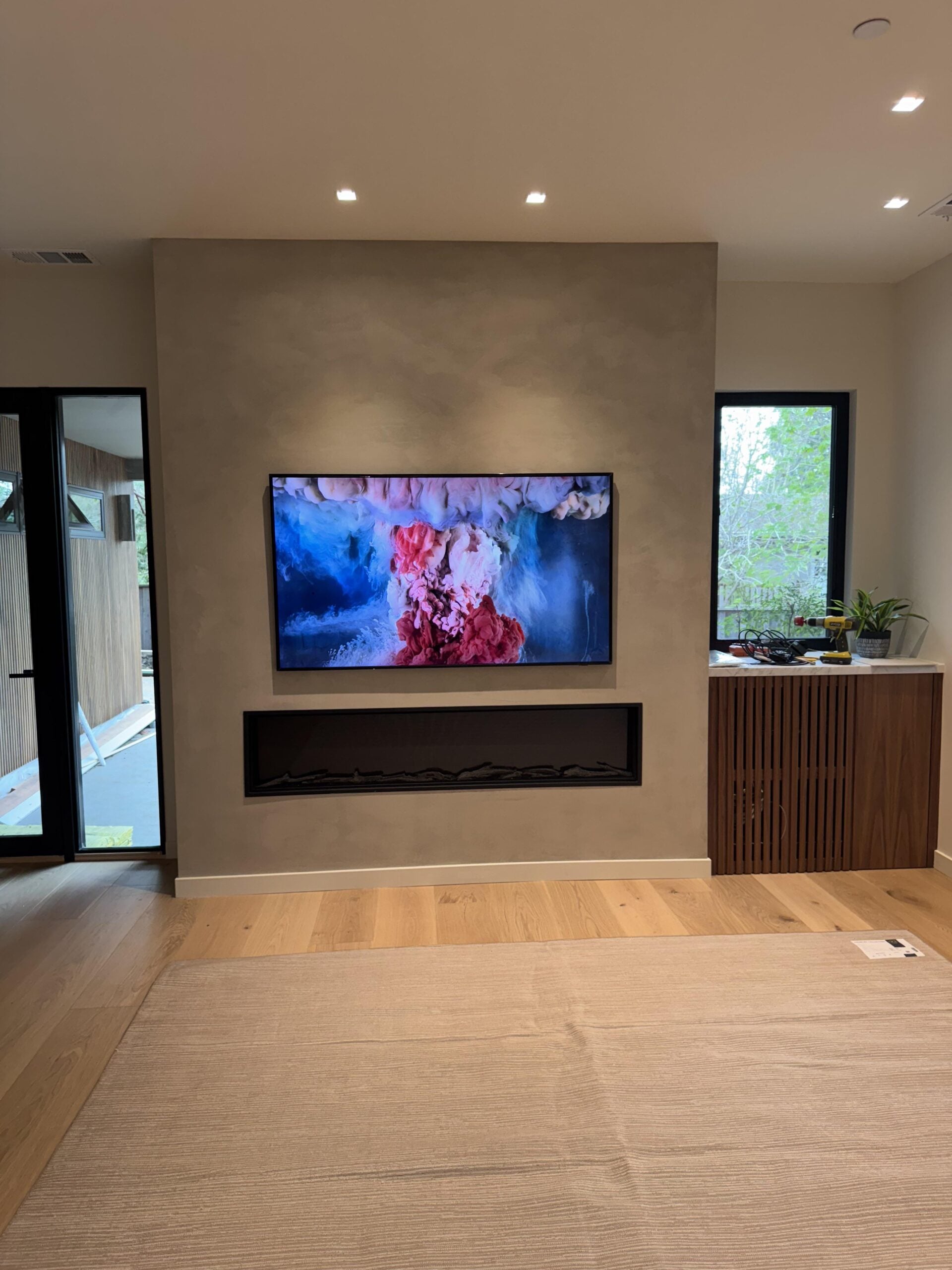 A wall-mounted TV displays colorful abstract art above a modern, recessed fireplace in a bright living room with wooden floors and a cabinet topped with plants and decor.
