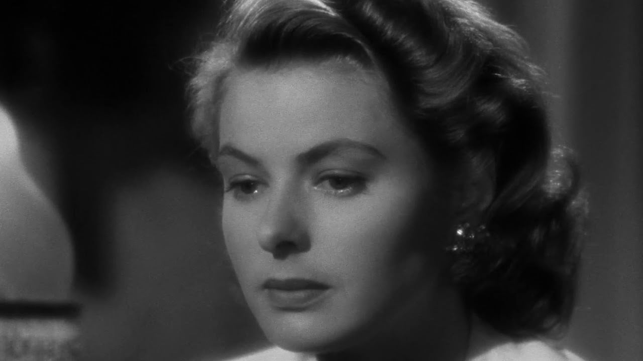 A woman with wavy hair looks down thoughtfully in a black-and-white close-up. Her expression appears serious and contemplative, with soft lighting highlighting her features.
