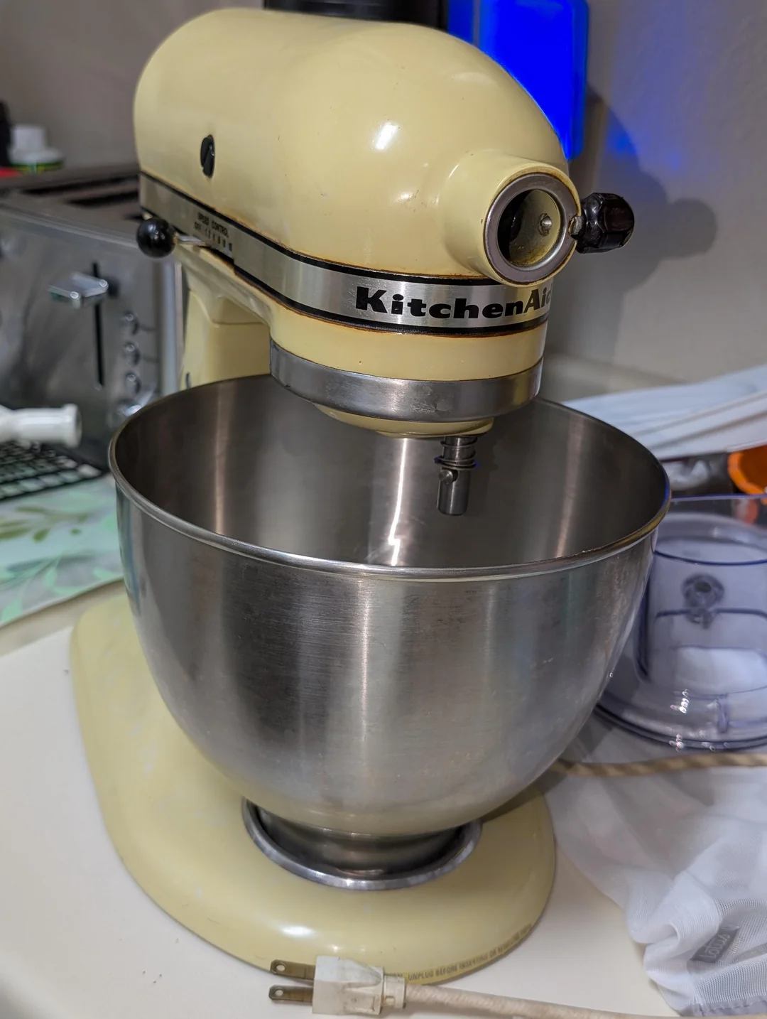A vintage cream-colored KitchenAid stand mixer with a stainless steel bowl sits on a kitchen counter next to a toaster and various utensils.