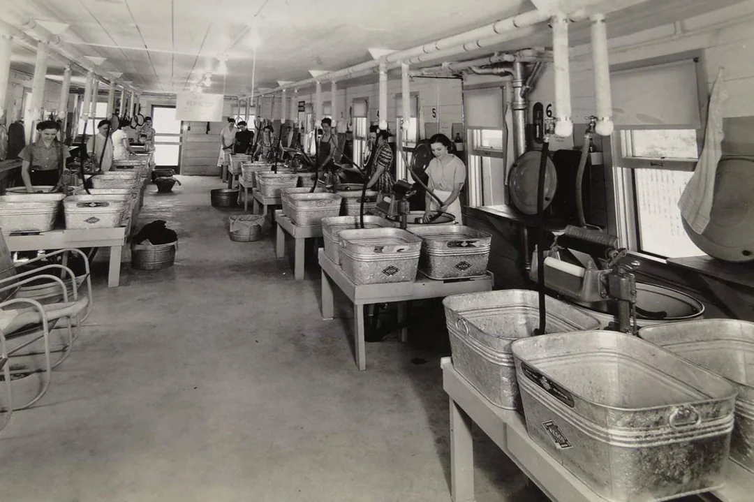 Black and white photo of women working in a large laundry room with rows of metal tubs and wringer washing machines. The room is long, with windows on one side and laundry equipment neatly arranged in rows.