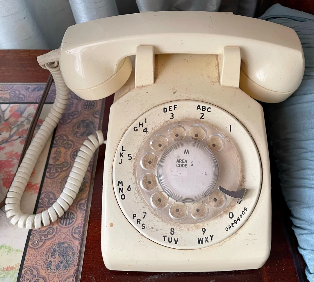 A beige rotary dial telephone with a coiled cord sits on a wooden surface near a blue cushion and a decorative mat. The phone shows some dust and signs of age.