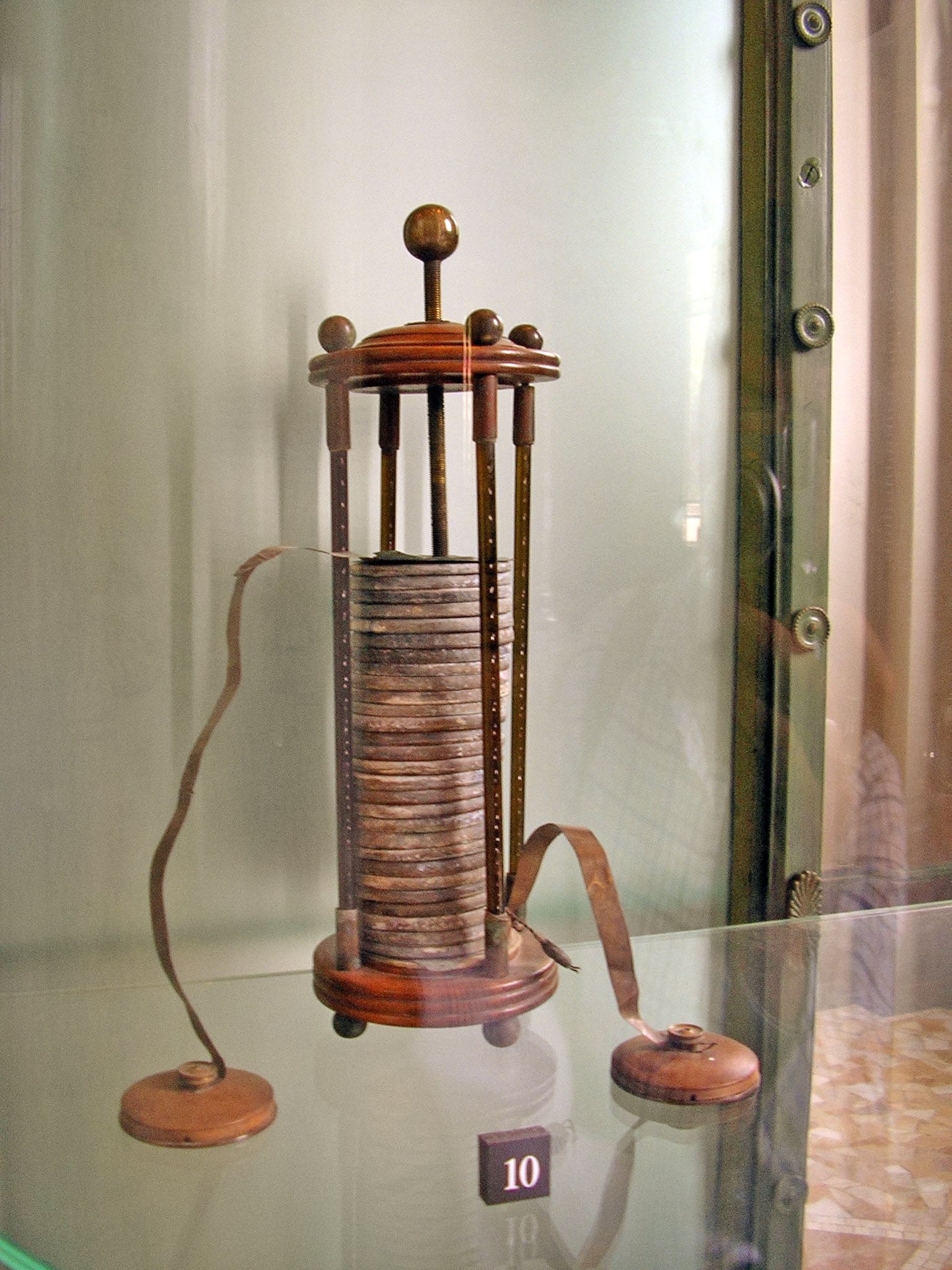A historic scientific instrument with a cylindrical stack of metal discs, wooden supports, and ribbon-like connectors, displayed in a glass case.