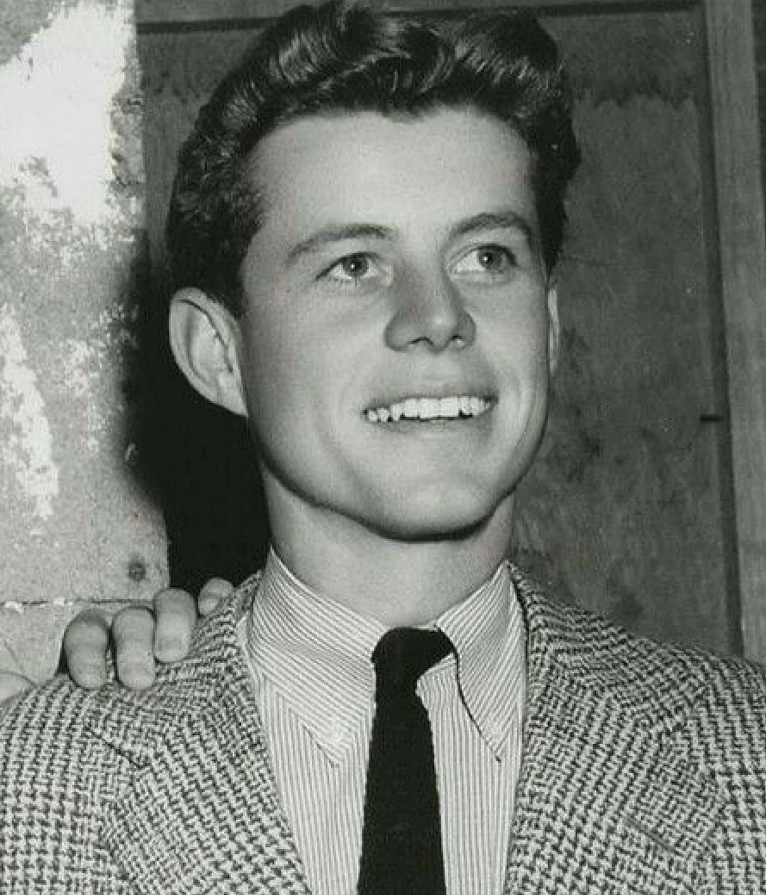 A young man in a checkered jacket, striped shirt, and dark tie smiles while looking slightly to the side. A hand rests on his shoulder. The black-and-white photo has a textured wall in the background.