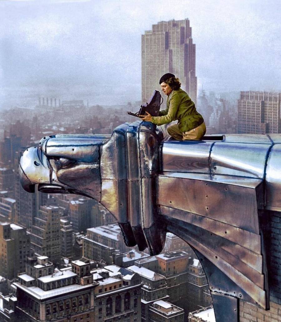 A person kneels on the edge of a tall building’s metallic eagle gargoyle, setting up a camera, high above a cityscape with snowy rooftops and skyscrapers in the background.