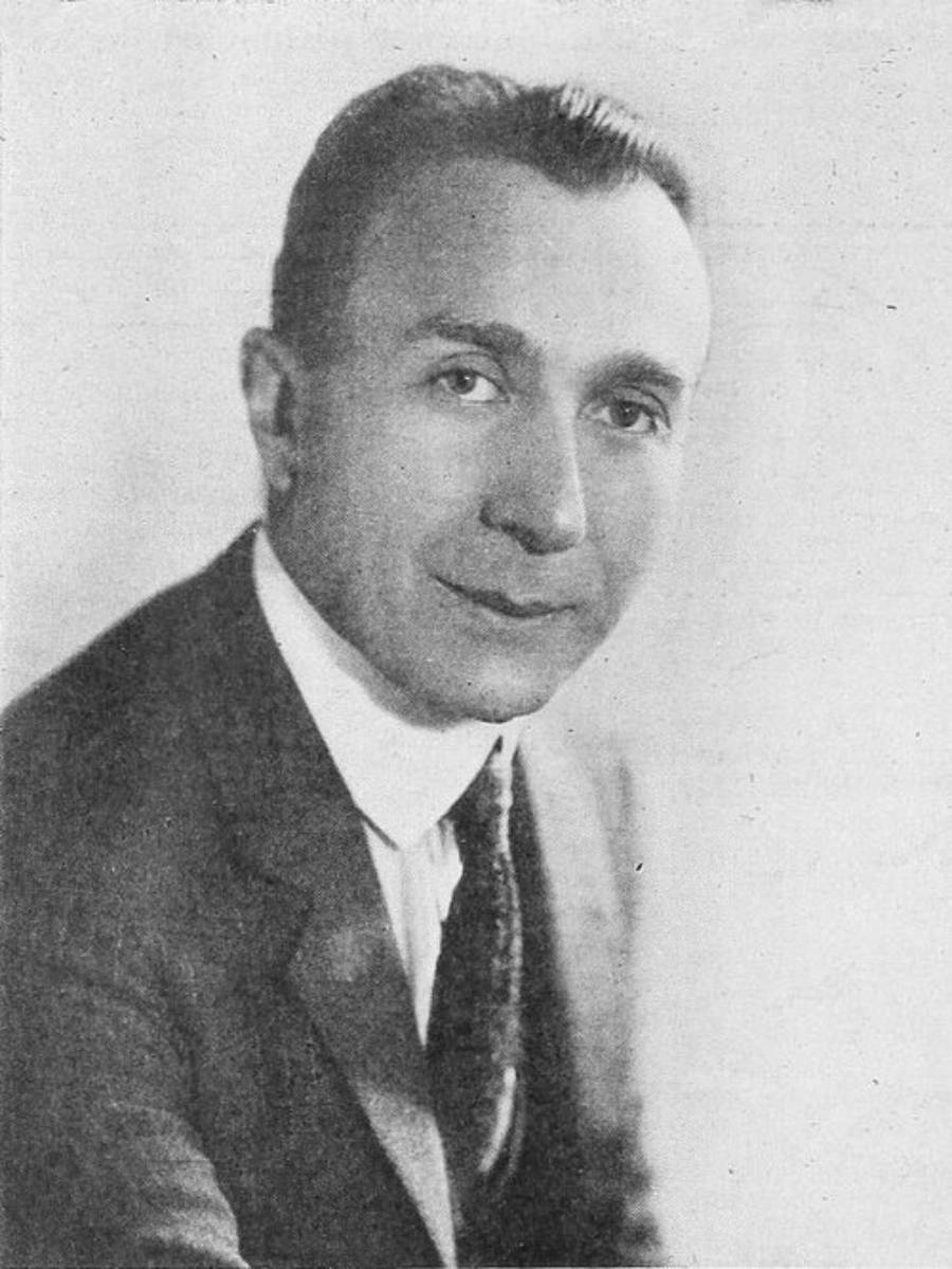 Black-and-white portrait of a man in a suit and tie, looking at the camera and smiling slightly, with short, neatly combed hair, on a plain background.
