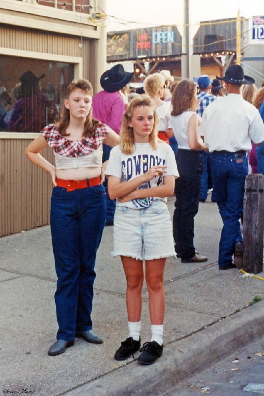 Two young women stand on a sidewalk. One wears a plaid shirt, high-waisted jeans, and a red belt. The other wears a “Cowboys” tee, denim shorts, and black shoes. People and a cowboy-themed setting are in the background.