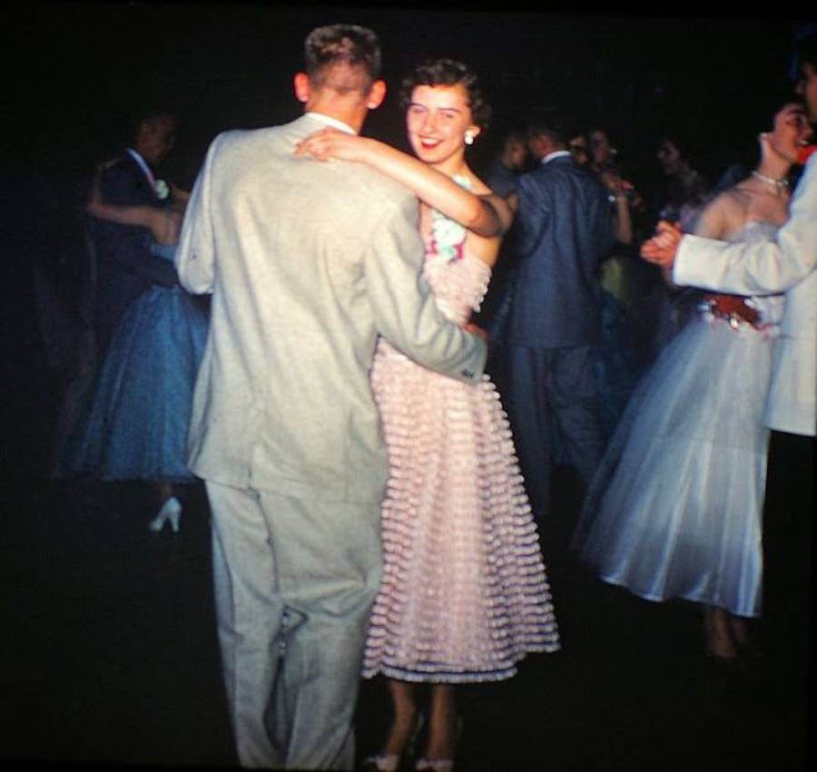 A young couple dances at a formal event; the woman wears a light pink dress and smiles at the camera, while others dance in the background, dressed in 1950s-style attire.