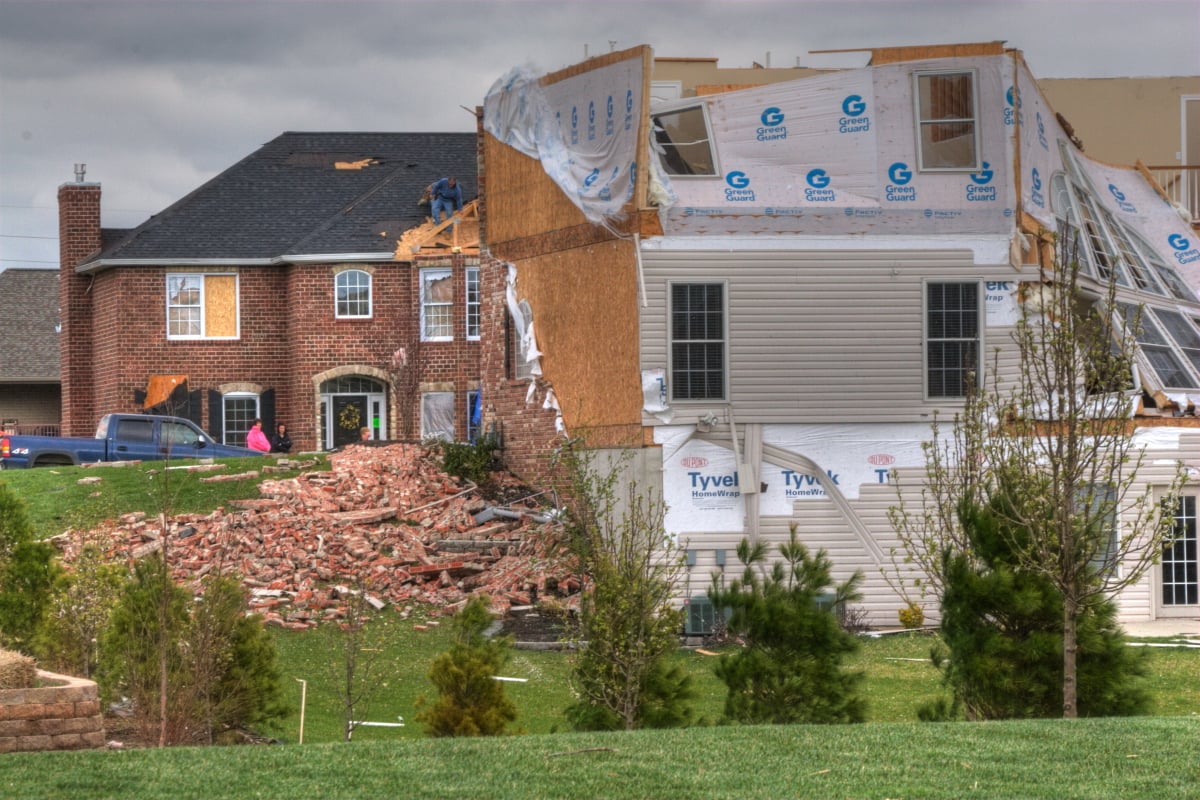 A partially destroyed house with debris scattered outside stands beside an intact brick home. People are on the roof of the intact house, working. The sky is cloudy and the scene suggests recent storm damage.