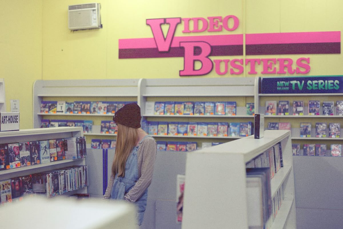 A person in a striped shirt and beanie browses movies in a retro video rental store with a large “Video Busters” sign and shelves of DVDs and VHS tapes.