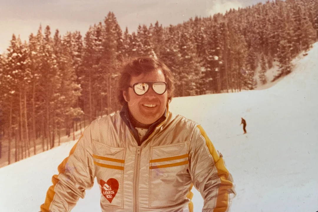 A man in a shiny silver jacket with yellow stripes smiles while standing on a snowy slope, wearing reflective sunglasses. Pine trees and a skier are visible in the background.