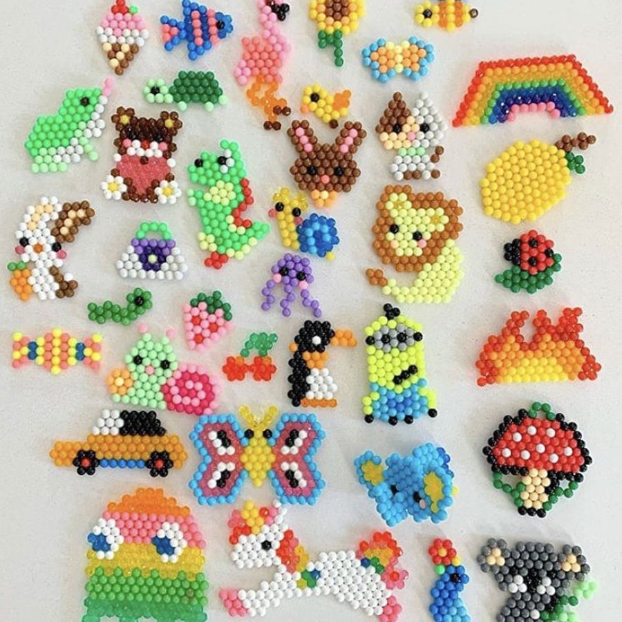 Colorful bead art mosaics on a white surface, featuring various shapes like animals, a rainbow, a butterfly, a unicorn, fruit, and objects such as a car and a mushroom, all arranged in a scattered, playful layout.