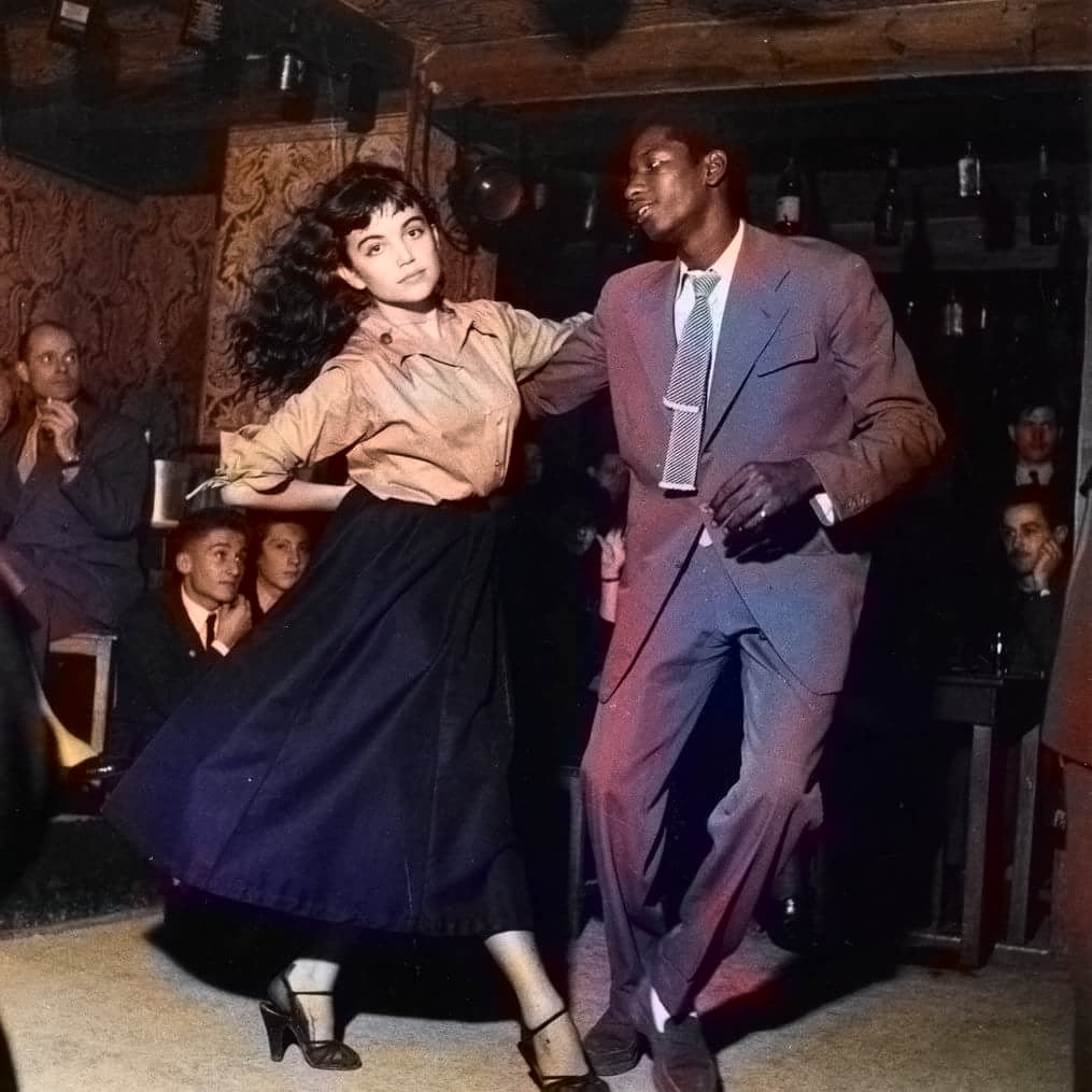 A woman and a man dance energetically together in a crowded club, both dressed formally, with onlookers seated nearby watching them.