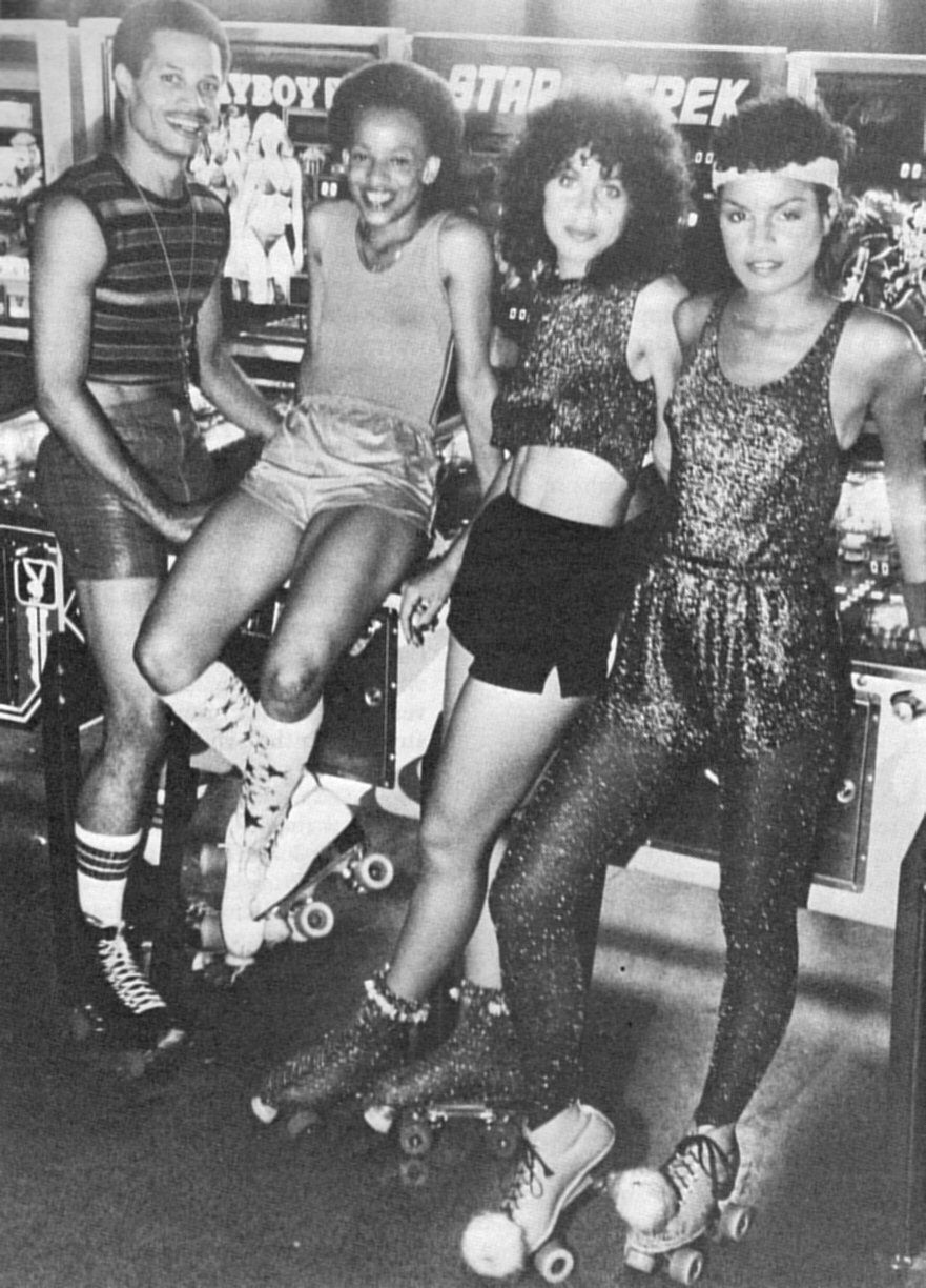 Four people in roller skates pose together in front of arcade game machines. They wear retro, sparkly outfits and smile at the camera.