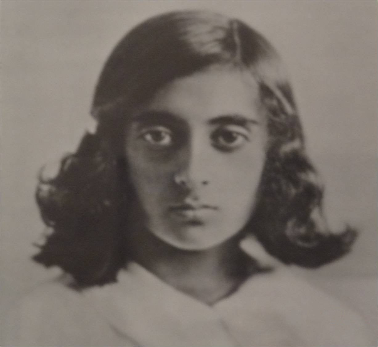 A black and white portrait of a young person with shoulder-length dark hair, looking directly at the camera with a neutral expression, wearing a light-colored top.