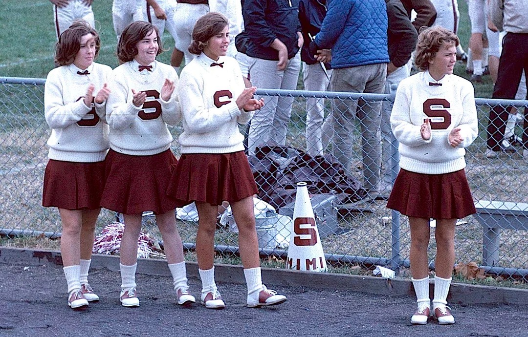 Four cheerleaders in matching sweaters with the letter “S” and maroon skirts stand and clap by a fence at a sports field. People and a cone with an “S” are visible in the background.