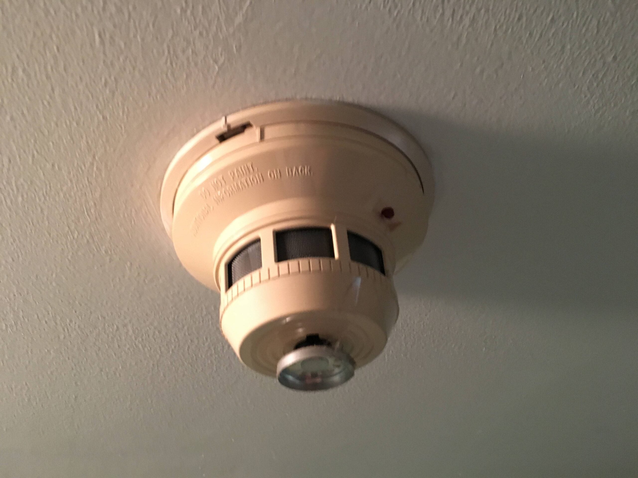 A beige smoke detector is mounted on a white ceiling, showing a small red indicator light and a clear plastic dome in the center.