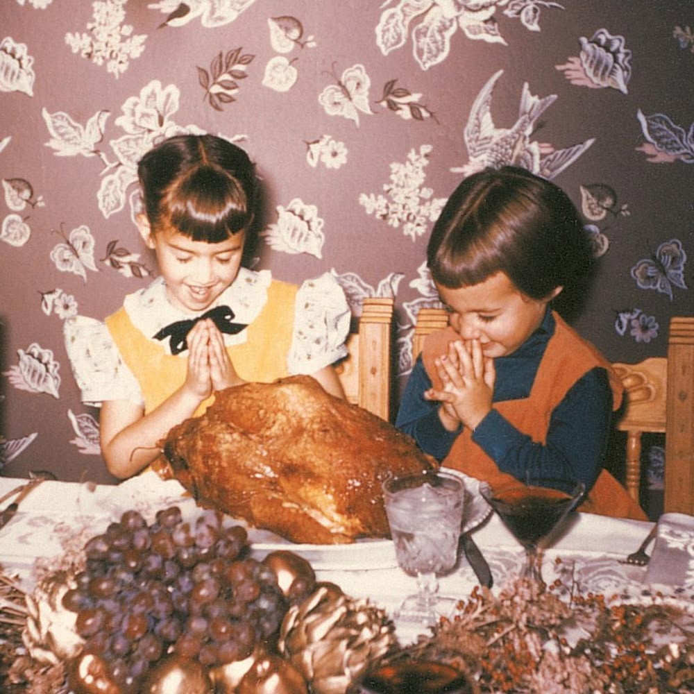 Two young girls sit at a table, hands clasped in prayer, smiling at a large roast turkey. The table is set with food and drinks. The background has wallpaper with bird and plant designs.