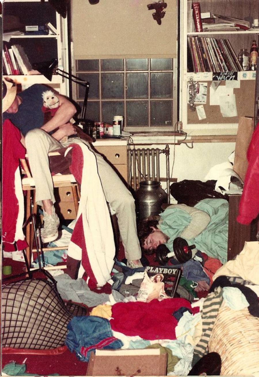 A cluttered, messy room with clothes, books, and objects scattered. Two people are in the room, one seated with a puppet, and one lying on the floor near a magazine. Shelves, a lamp, and a window are visible in the background.