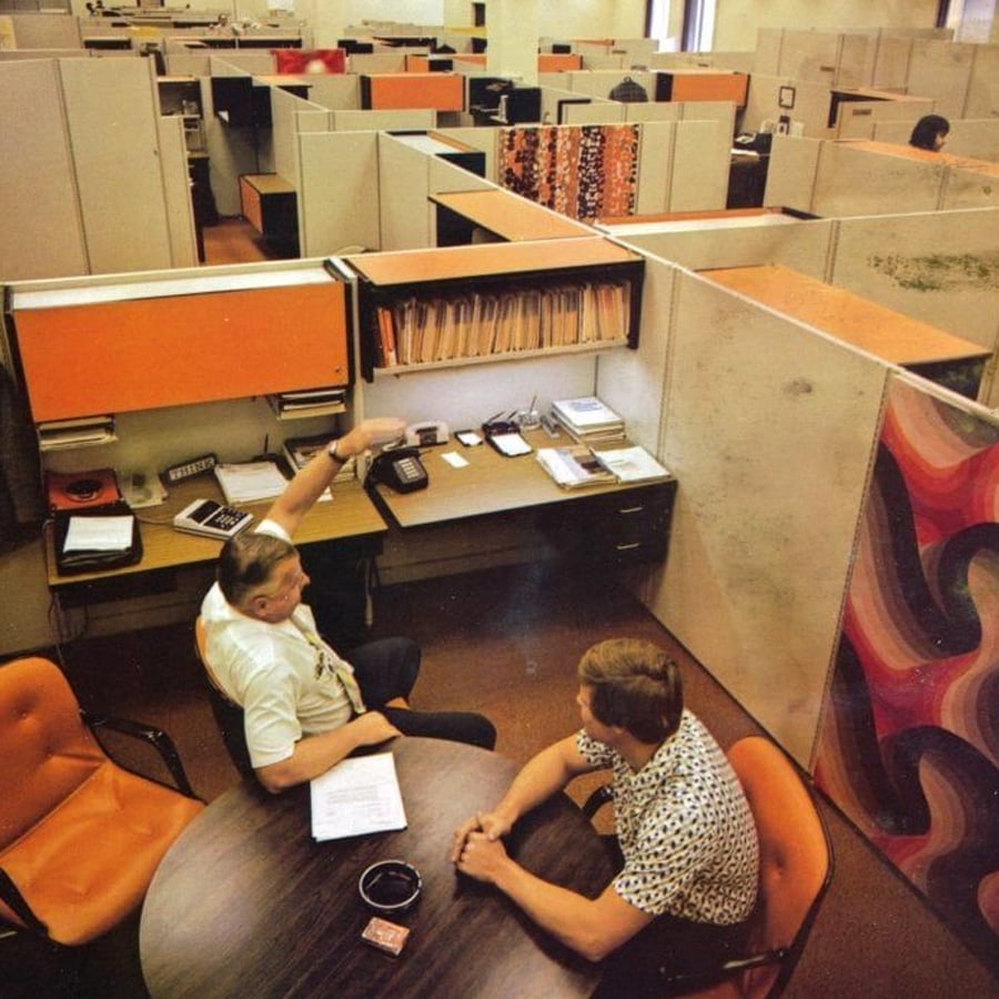 Two men sit at a round table in a retro office with orange chairs and beige cubicles. One gestures while talking. Files, phones, and papers are on the desk; patterned curtains hang in the background.