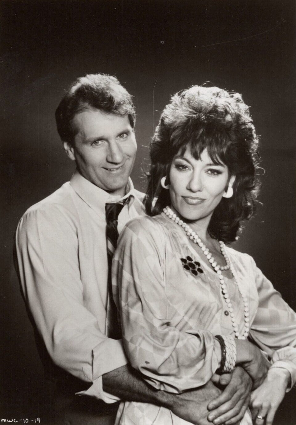 A man in a shirt and tie stands behind a woman in a dress and pearls, both smiling and embracing against a dark studio background.