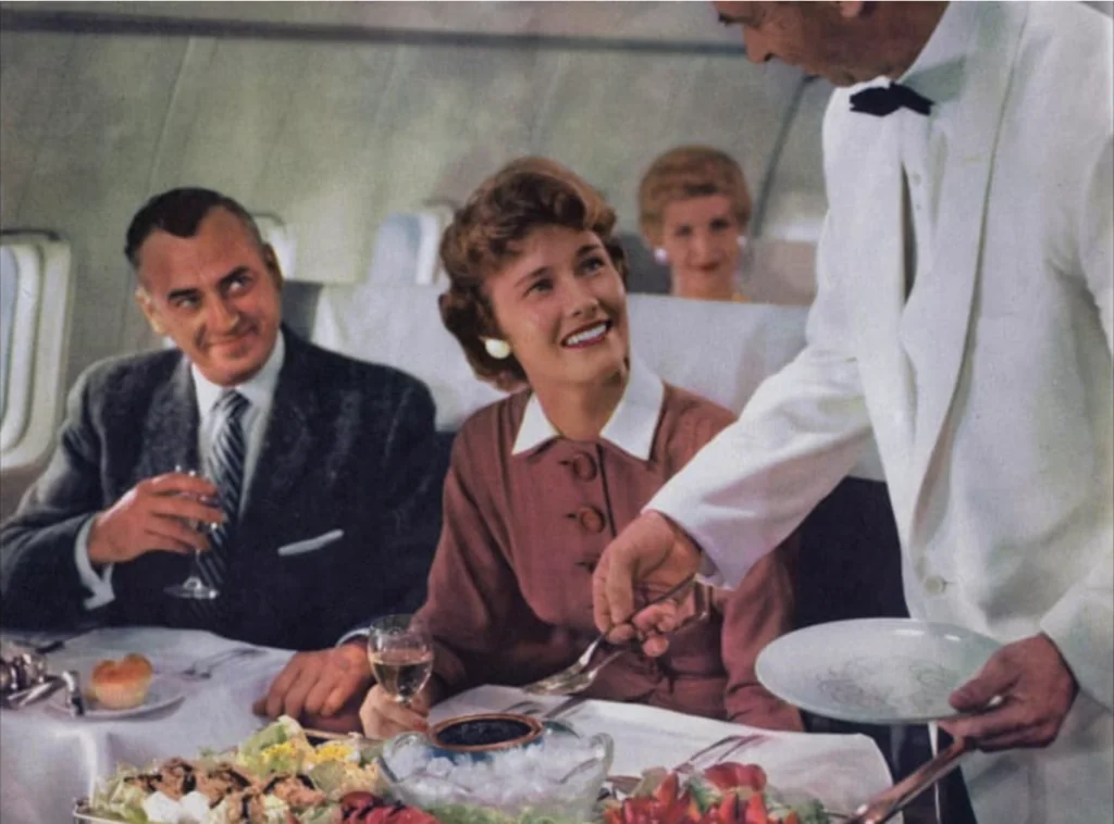 A flight attendant in a white uniform serves a smiling woman and a man at a table set with food and drinks inside an airplane. Another woman sits in the background.