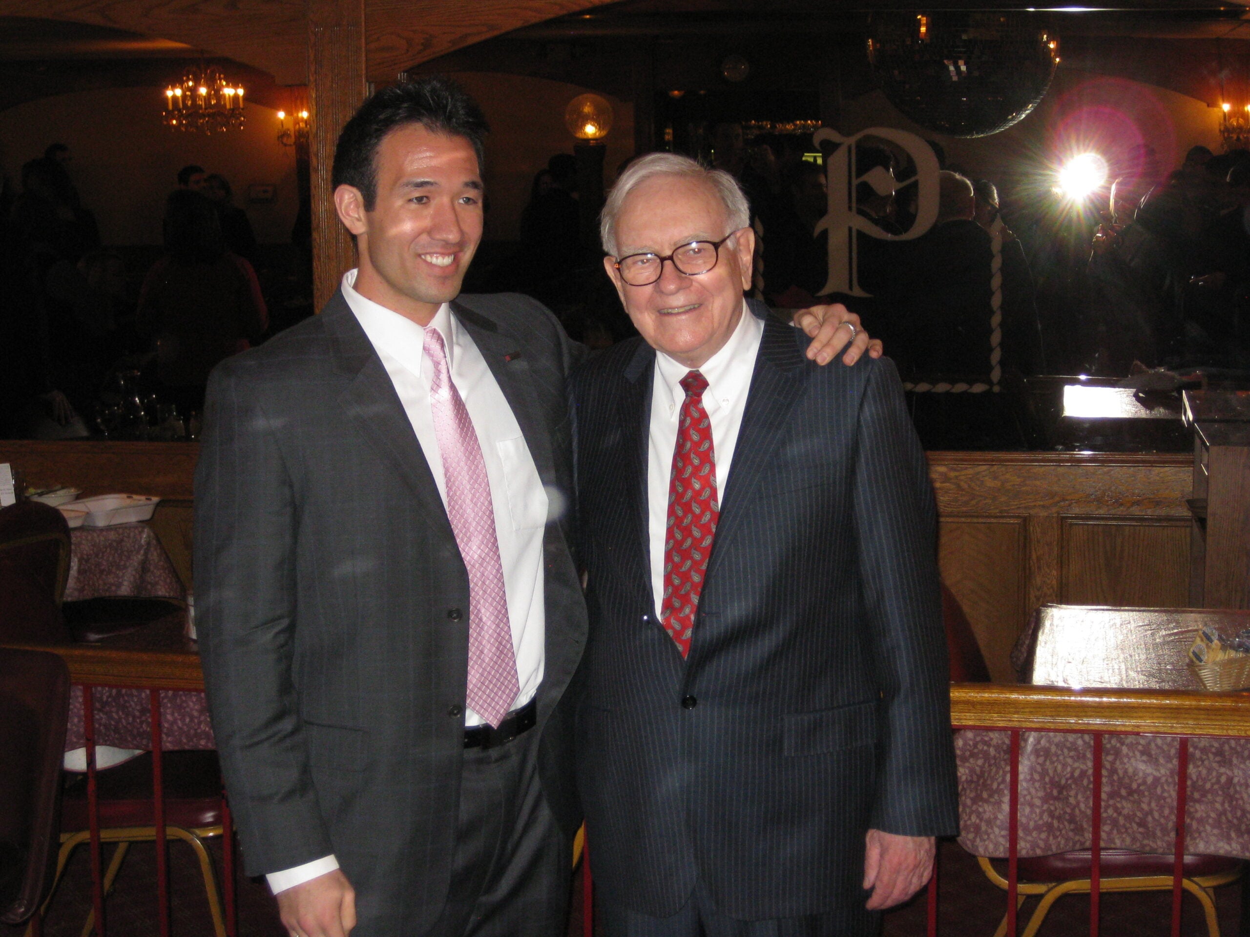 Two men in suits stand together and smile at an indoor event. One has his arm around the other. There are chairs, tables, and people in the dimly lit background.