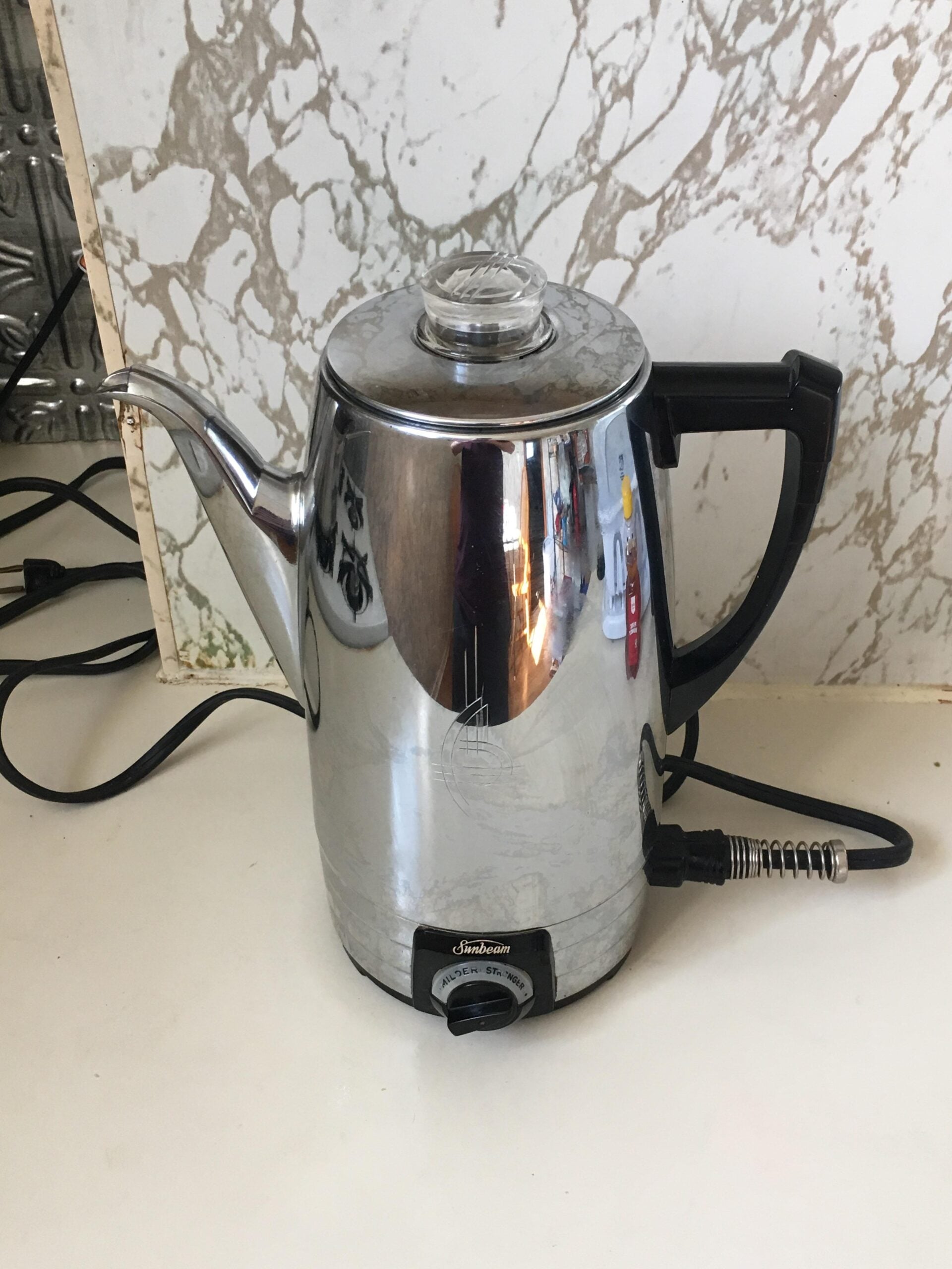 A stainless steel electric percolator with a black handle sits on a kitchen counter against a marble-patterned backsplash. The power cord is plugged in, and the percolator’s surface reflects nearby objects.