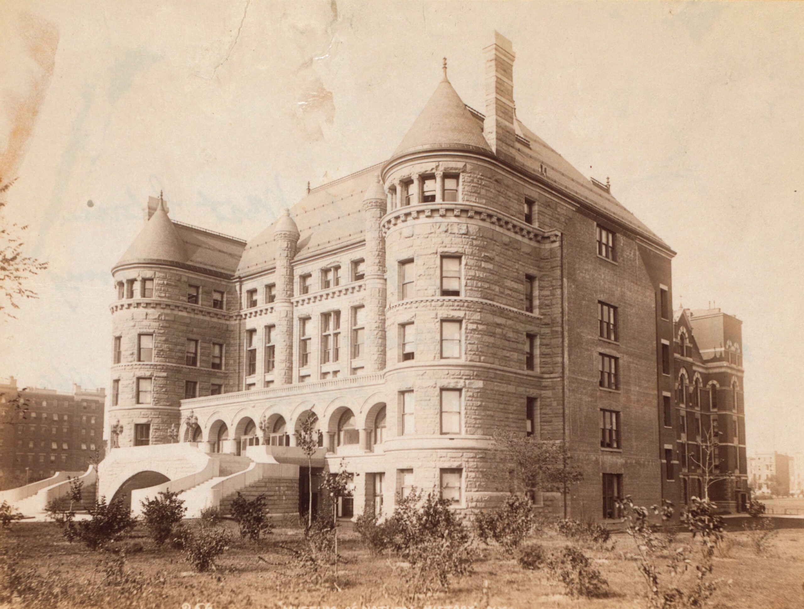 Sepia photo of a large, historic stone building with rounded towers, arched windows, and a grand staircase in front, surrounded by grass and small trees, set in an urban area.