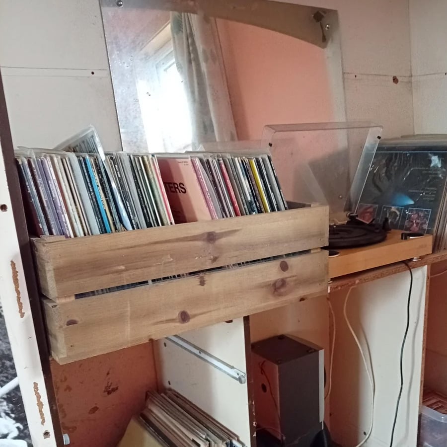 A wooden crate filled with vinyl records sits on a shelf below a mirror. A record player, speakers, and more records are nearby in a cozy, cluttered room with warm lighting.