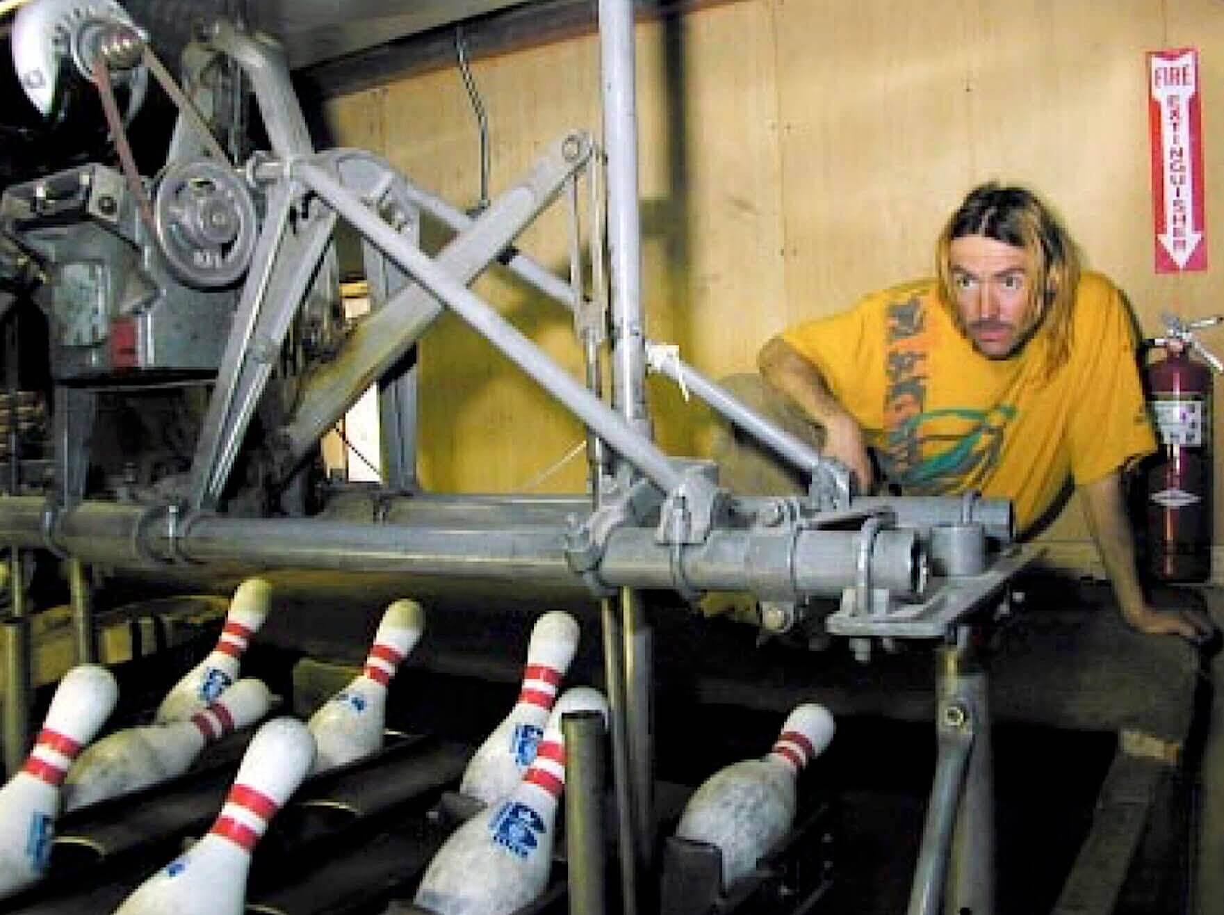 A person with long hair and a yellow shirt inspects a mechanical bowling pinsetter beside a row of standing pins. A fire extinguisher is mounted on the wall nearby.