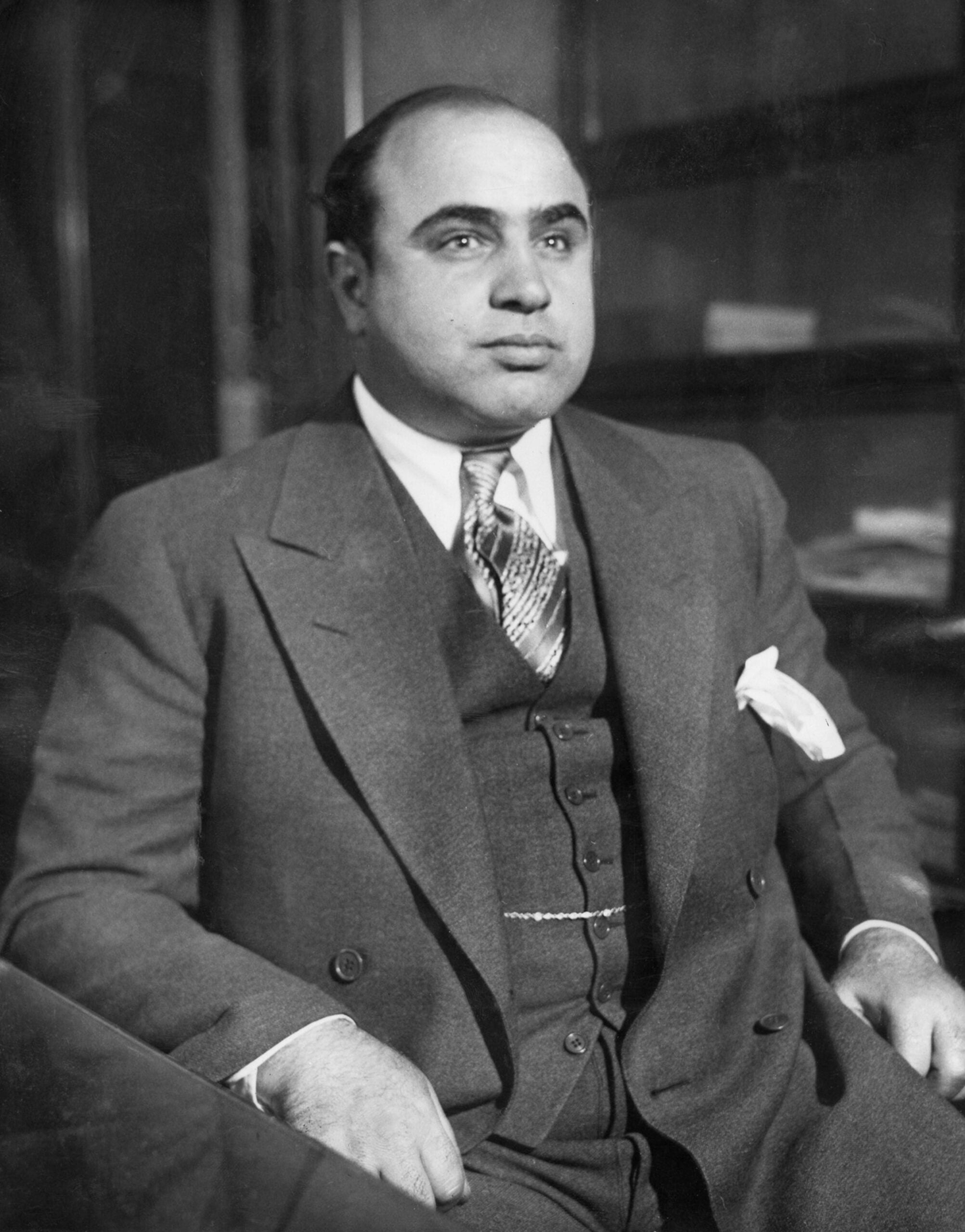 A man in a suit and tie sits at a desk, looking slightly to the side. He has short dark hair and a serious expression. The background is dark and blurred.