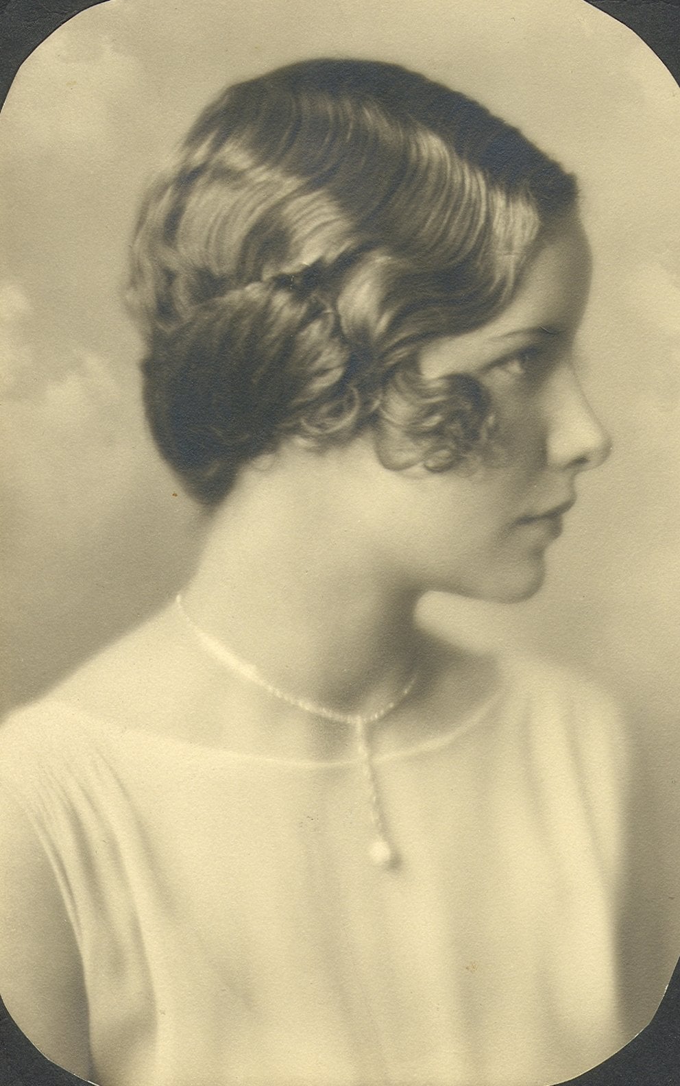A vintage sepia portrait of a young woman in profile, with short, wavy hair and a beaded necklace with a pendant, wearing a light, sheer top.