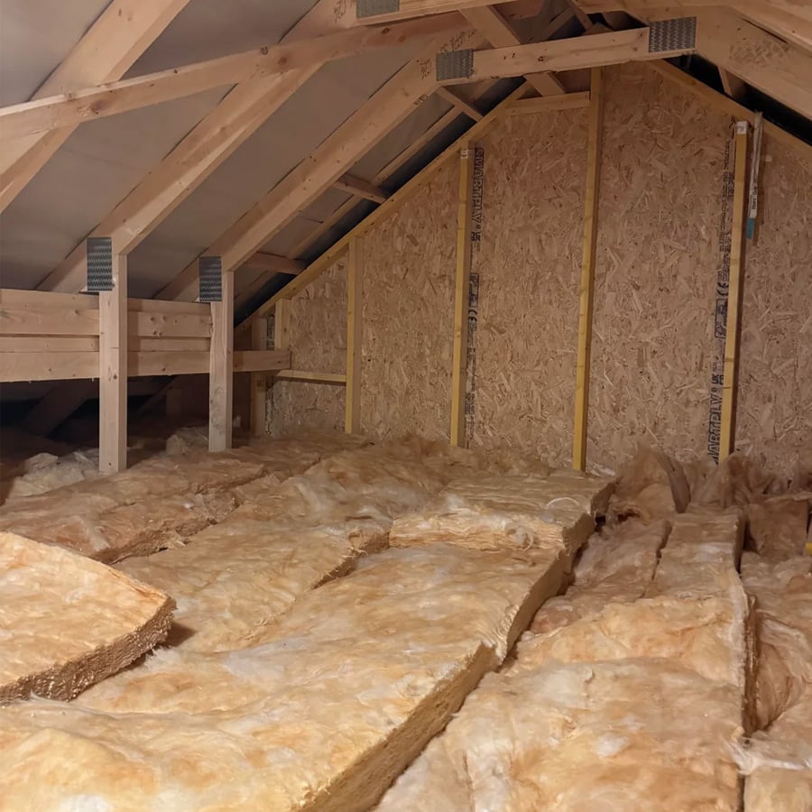 Attic space with wooden beams and yellow fiberglass insulation laid on the floor, with unfinished plywood walls visible in the background.