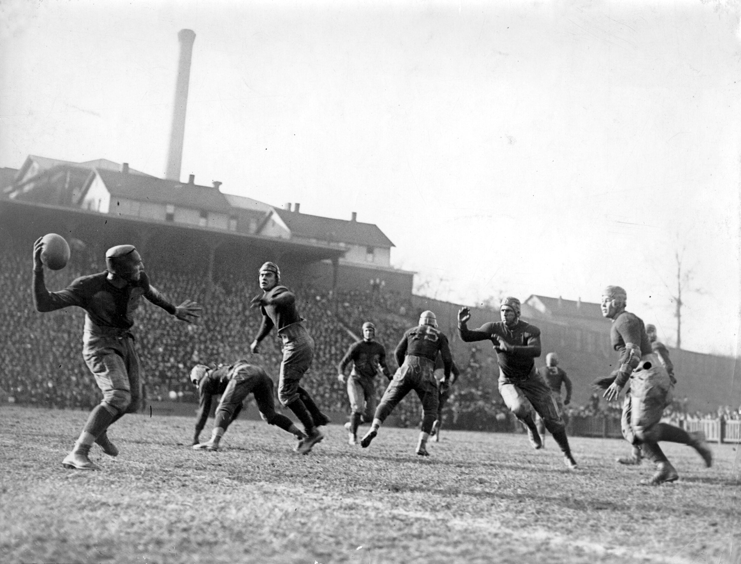 Seven American football players compete on a grassy field in an old black-and-white photo, with a large crowd and buildings visible in the background. One player is preparing to throw the ball.