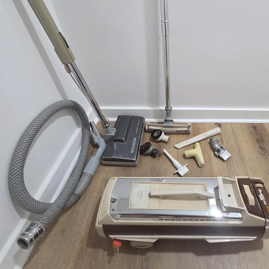 Vintage canister vacuum cleaner with a gray hose, several metal and plastic attachments, and a floor head, all placed on a wood floor against a white wall.