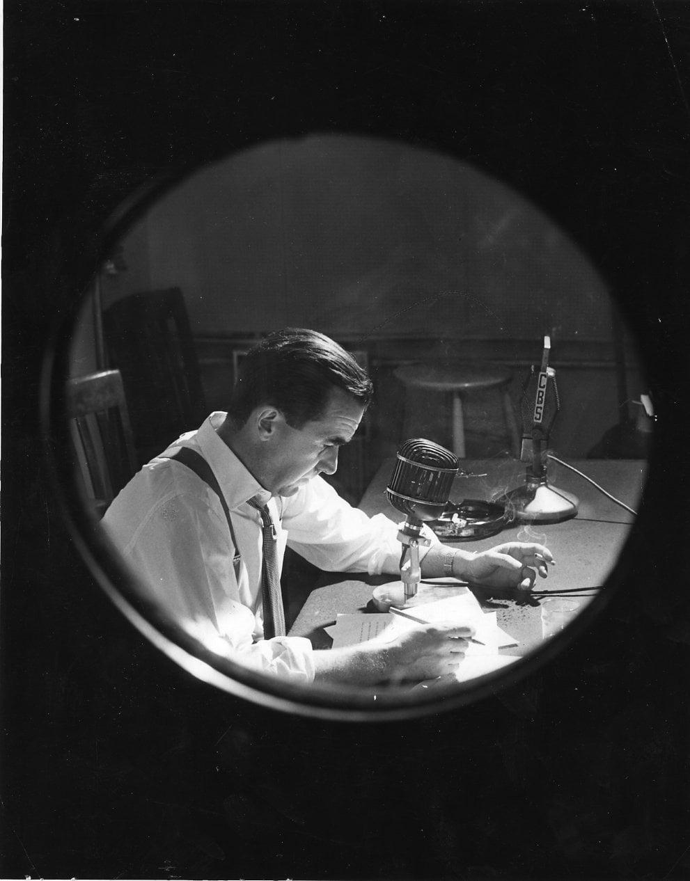 A man sits at a desk in front of a microphone, reading papers, as seen through a circular window or lens. The scene appears to be from a radio broadcast studio.