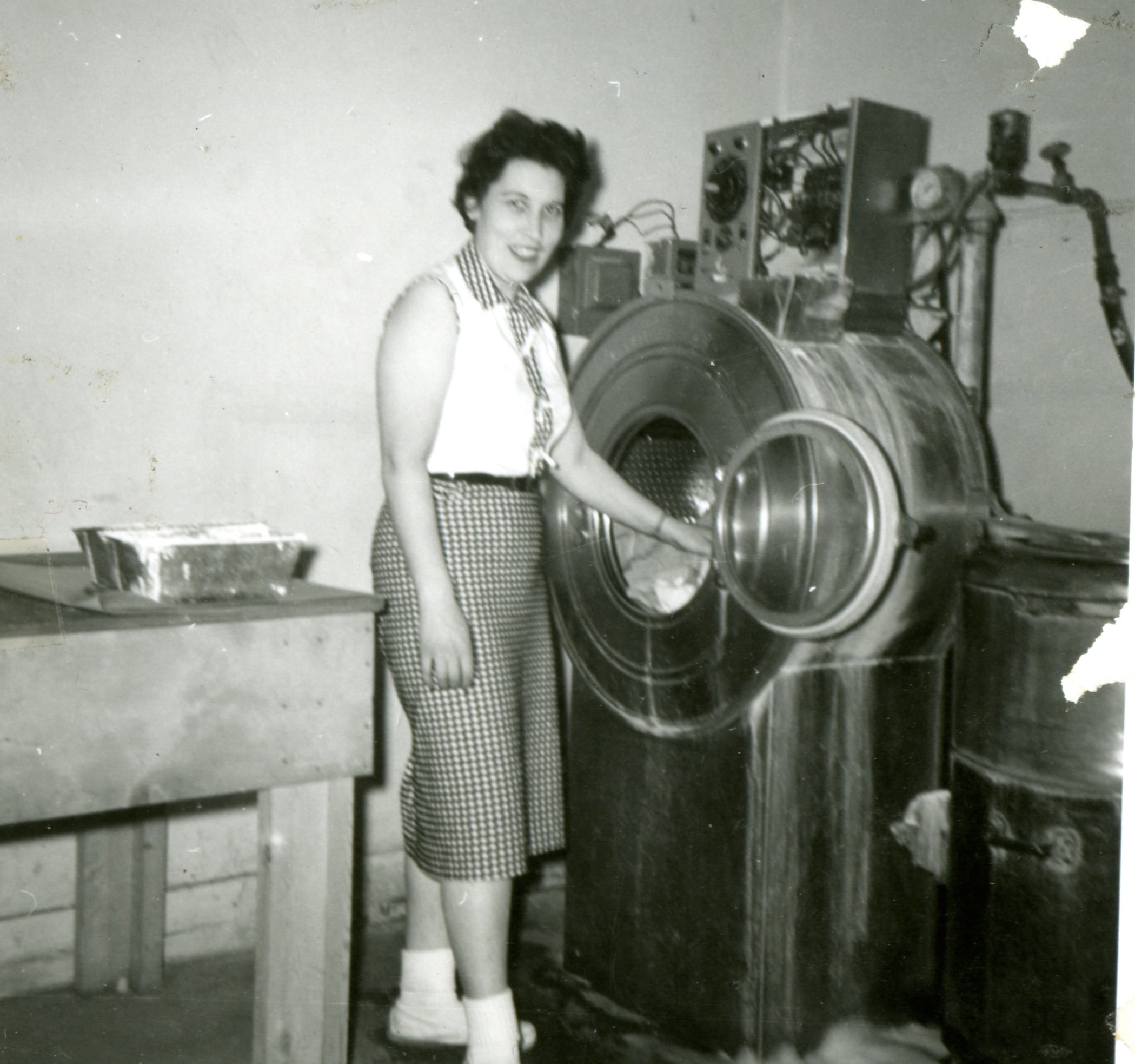 A woman stands beside a large, open-front industrial washing machine in a laundry room, smiling at the camera. She wears a sleeveless blouse, checked skirt, socks, and shoes.
