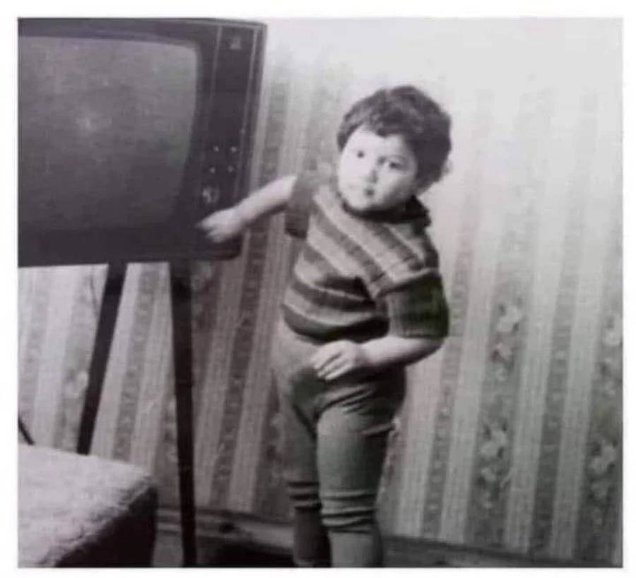 A small child in striped clothes stands next to an old-fashioned television, reaching to touch its knobs. The room has patterned wallpaper and a chair is partially visible in the foreground. The photo is black and white.