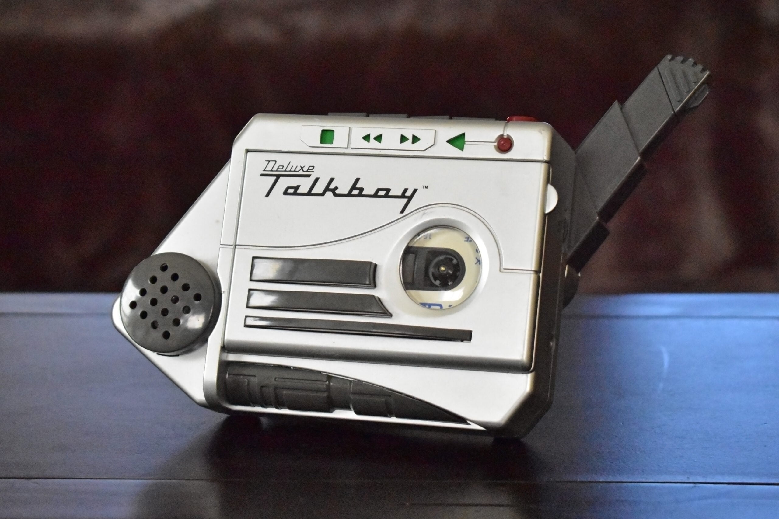 A silver Deluxe Talkboy cassette recorder sits on a dark surface with its microphone extended, showing buttons and a cassette window.